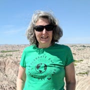 A smiling white woman with shoulder-length grey hair in sunglasses and a green Earth Day tshirt in front of a rocky canyon landscape. 