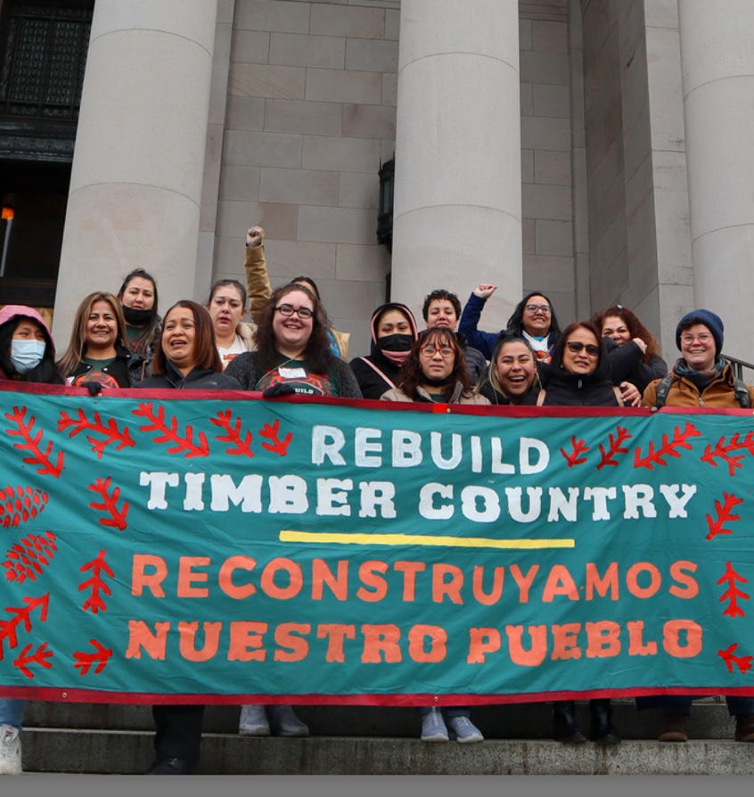 A group of women smile in front of a building with pillars, holding up a turquoise sign with a red border that reads "Rebuild Timber Country / Reconstruyamos Nuestro Pueblo' 