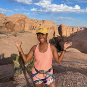 Kristen Walker, in a peach hiking tank and patterend shorts, stands in front of a desert landscape with large boulders and blue sky with her hands raised to the landscape behind her. 